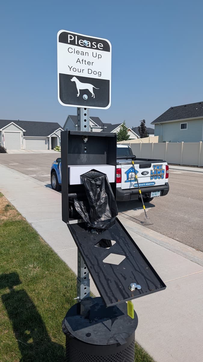 A Lawn Dogs technician's truck beside a pet waste station with its bin open and the liner being replaced, under a Please Clean Up After Your Dog sign